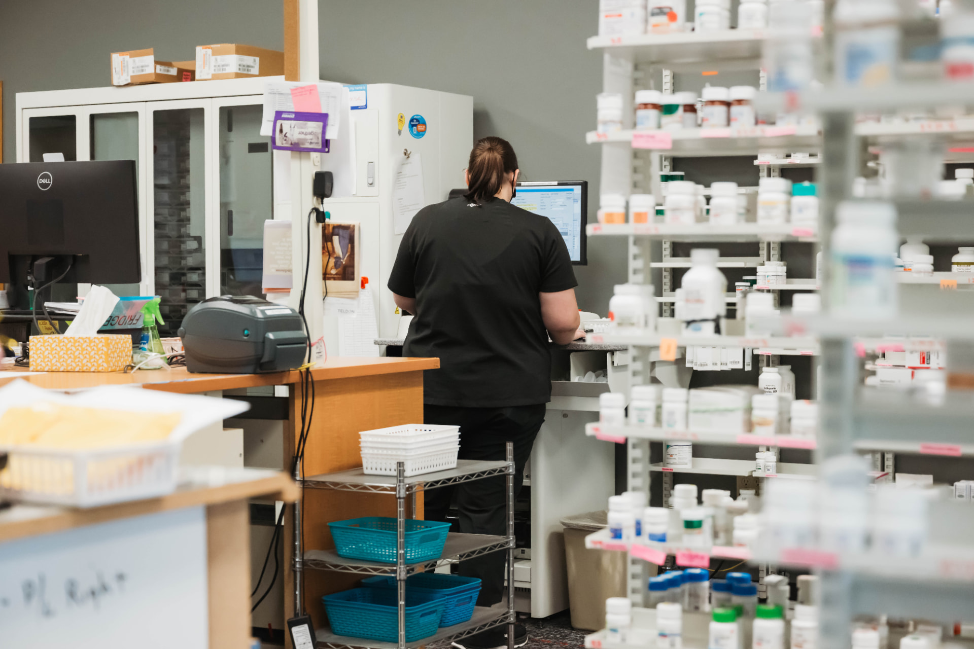 Pharmacy staff filling prescriptions at a workstation beside stocked medication shelves